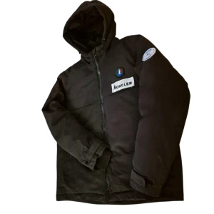 Svart Moncler pufferjacka med huva - Svart pufferjacka från Moncler Grenoble med huva och dragkedja. Jackan har två bröstfickor, patchar på bröstet och ärmen samt diskret Moncler-logga. Materialet är syntet och jackan är designad för hög prestanda och intensiv sport. Perfekt för kalla dagar.