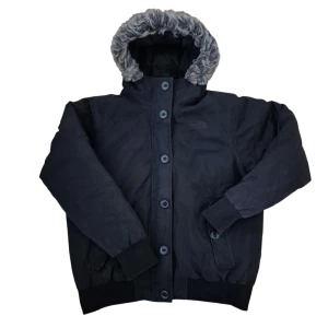 Svart pufferjacka från The North Face bomber  - Svart pufferjacka från The North Face med fluffig grå fuskpäls runt huvan. Jackan har knappar framtill, ribbade muddar och två fickor. Insidan är quiltad för extra värme och huvan är stor och mysig. Perfekt för kalla vinterdagar.