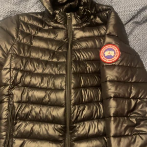 Canada goose puffer jacka - Säljer en svart pufferjacka från Canada Goose med huva och klassisk logga på ärmen. Jackan har quiltad design, dragkedja framtill och är perfekt för kalla dagar. Materialet är syntet och dun, vilket gör den både lätt och varm. Passformen är normal och jackan har långa ärmar. Köpt tidigt detta året, köpt en ny jacka då därför den säljs, Kvitto medföljer (skickas i dm) vid en snabbaffär fixar vi ett riktigt bra pris, ny pris 10.000kr