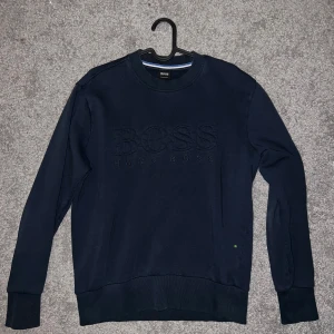 Mörkblå sweatshirt från Hugo Boss - Snygg mörkblå sweatshirt från Hugo Boss med broderad logga framtill. Tröjan har rund halsringning, långa ärmar och ribbade muddar vid ärmslut och nederkant. Perfekt för en stilren och avslappnad look.