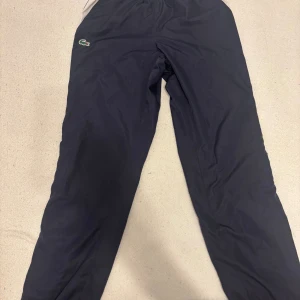 Mörkblå träningsbyxor Lacoste Sport - Snygga mörkblå träningsbyxor från Lacoste Sport med vita och svarta rutiga sidopaneler och orange detaljer. Byxorna har elastisk midja och regular fit, perfekt för sportiga dagar. Klassisk Lacoste-logga på benet.