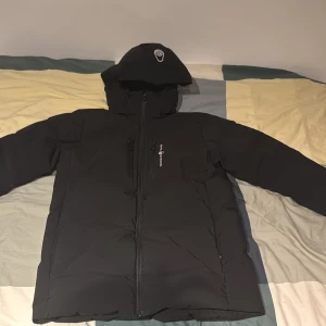 Svart dunjacka Sail Racing - Svart dunjacka från Sail Racing med huva och snygga logodetaljer på bröstet och ärmen. Jackan har dragkedja framtill, flera fickor och är fodrad med dun för extra värme. Perfekt för kalla dagar och har en clean, sportig look.