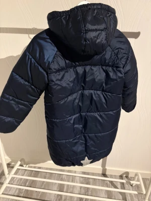 Mörkblå pufferjacka från Lindex - Säljer en mörkblå pufferjacka från Lindex med huva och mysigt ljust foder. Jackan har långa ärmar, två fickor med knapp och en snygg dragkedja i kopparfärg. Perfekt för kalla dagar och har en cool quiltad look.