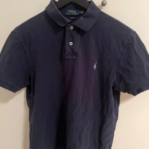 Mörkblå piké från Polo Ralph Lauren - Snygg mörkblå pikétröja från Polo Ralph Lauren med klassisk krage och två knappar framtill. Tröjan har korta ärmar och den ikoniska broderade loggan på bröstet. Tillverkad i mjuk bomull som är skön mot huden.
