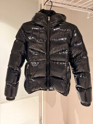 Svart jacka Colmar - Säljer en svart, glansig pufferjacka från Colmar med huva och två sidofickor med dragkedja. Jackan har ett stilrent, quiltat mönster och logotyp på ärmen. Perfekt för kalla dagar och har en modern, loose passform. Inköpt förra säsongen. Sparsamt använd. Inget att anmärka. Kvitto finns. 