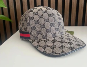 Gucci keps med GG-mönster och röd-blå detalj - Snygg keps från Gucci med klassiskt GG-mönster i mörkgrått på beige bakgrund. Kepsen har en böjd skärm och en röd-blå rand på sidan. Justerbar rem baktill i svart skinn. Perfekt för dig som vill sticka ut med lyxig streetstyle.