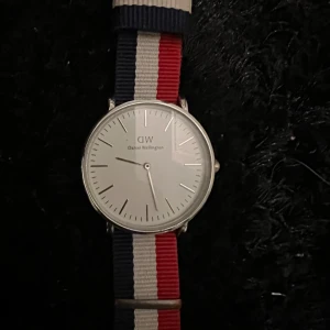 Randig klocka från Daniel Wellington - Snygg klocka från Daniel Wellington med rund silverfärgad boett och vit urtavla. Armbandet är randigt i blått, vitt och rött och ger en cool och ungdomlig vibe. Perfekt accessoar för dig som vill sticka ut med stil. I jättefint skick, behöver endast batteribyte då jag inte anvönt den på länge.
