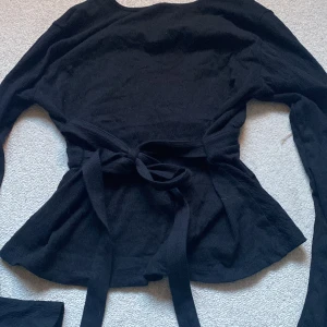 Svart omlottopp med knyt från Gina Tricot - Svart omlottopp från Gina Tricot i storlek XS. Toppen har lång ärm, v-ringad hals och ett brett knytband i midjan som ger en snygg siluett. Materialet är ribbat och mjukt, perfekt för att skapa en cool look.