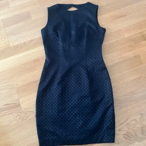 Svart mönstrad kort klänning H&M - Snygg svart klänning från H&M med diskret geometriskt mönster. Modellen är ärmlös och har en tight passform med dragkedja i ryggen och en liten öppning med knapp i nacken. Materialet är polyester och klänningen är fodrad för extra komfort.