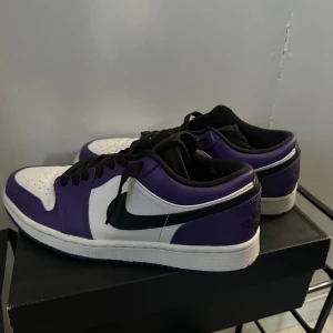 Nike Air Jordan 1 Low Court Purple - Nike Air Jordan 1 Low Court Purple med vit och lila ovandel i skinn, svart swoosh och svarta snören. Klassisk rund tå och platt sula, med perforeringar på tån för extra ventilation. Ikonisk Jordan-logga på hälen och röd innersula för en snygg kontrast.