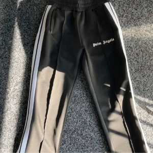 Palm Angels svarta mjukisbyxor med vita ränder - Svarta mjukisbyxor från Palm Angels med vita sidoremsor och logotyp broderad på vänster ben. Byxorna har elastisk midja och är tillverkade i polyester för en skön och sportig känsla. Perfekt för en streetwear-look. Lite slitage på knäna i sömnen som är uppvikt men inget som syns!!