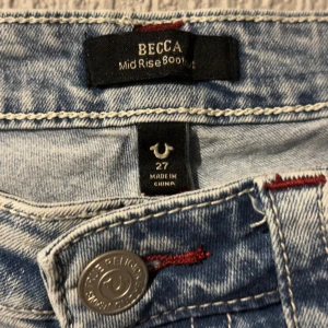 True Religion ljusa bootcut jeans - Snygga ljusblå jeans från True Religion, modell Becca Mid Rise Bootcut. Jeansen har slitna detaljer vid knäna, röda sömmar och klassiska fickor. Endast testade strl 27