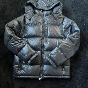 Svart dunjacka från Polo Ralph Lauren - Säljer en svart dunjacka från Polo Ralph Lauren med huva och dragkedja framtill. Jackan har en röd Polo-logga på bröstet, flera fickor med dragkedja och en glansig finish. Perfekt för kalla dagar och har en riktigt schysst puffer-look. Den är helt ny inga skador. Skriv vid funderingar