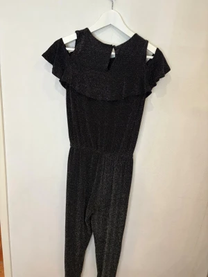 Glittrig svart jumpsuit med volang - Supersnygg svart jumpsuit från Kappahl med glittrigt tyg och volangdetalj över axlarna. Modellen har offshoulder och resår i midjan för en skön passform. Perfekt för dig som vill sticka ut med lite extra glitter och cool stil.