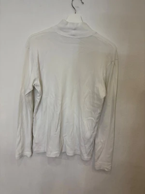 Vit polotröja från Simsoe  - Vit polotröja från Simsoe i mjuk bomull. Tröjan har lång ärm och en klassisk polokrage som ger en clean och stilren look. Perfekt att styla med jeans eller under en jacka när det är kyligt ute.