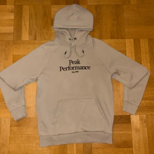 Beige hoodie från Peak Performance - Snygg beige hoodie från Peak Performance med svart logotyp på bröstet. Tröjan har huva med snörning, magficka och ribbade muddar vid ärmslut och nederkant. Perfekt för chill dagar och har en mjuk insida i fleece som gör den extra skön.