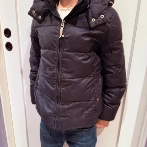 Mörkblå pufferjacka från GANT - Snygg mörkblå pufferjacka från GANT med huva och dragkedja framtill. Jackan har quiltad design, praktiska fickor och coola detaljer med tryckknappar på huvan. Perfekt för kalla dagar och har en klassisk look med GANT-logga på insidan. Tryckknapparna vid fickorna är lite skavda i färgen samt att det finns något väldigt litet hål som man ser på bild. 