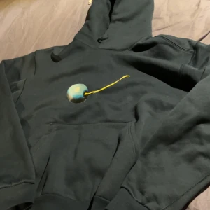 Mörkblå Nike SB hoodie med print - Svart hoodie från Nike SB med en stor ficka framtill och huva. På bröstet finns ett coolt print med en rund blågrön boll och gul linje. Liten Nike SB-logga på ärmen. Perfekt för dig som gillar streetwear och vill ha något unikt.