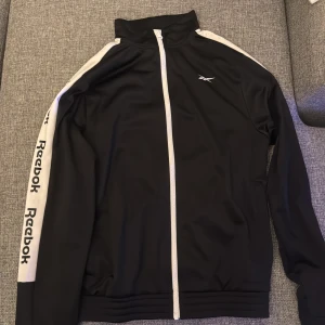 Svart kofta  - Svart kofta från Reebok i storlek S med vita detaljer och logga längs ärmarna. Jackan har hel dragkedja framtill, högt krage och långärmad design. Materialet är polyester och jackan har en sportig look med kontrastpaneler på axlar och ärmar.