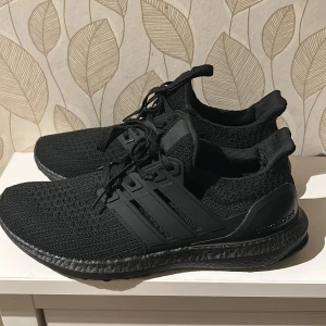 Svarta Adidas Ultraboost sneakers - Säljer ett par svarta Adidas Ultraboost sneakers med stickad ovandel och platt sula. Skorna har snörning och en snygg, sportig design med diskreta Adidas-loggor. Materialet är syntet och stickad textil för extra komfort och flexibilitet.