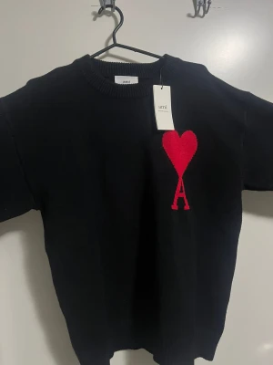 Svart stickad tröja från Ami Paris - Svart stickad tröja från Ami Paris med ikoniskt rött hjärta och A på bröstet. Klassisk rund halsringning, ribbade muddar och lång ärm. Perfekt för dig som gillar stilrena plagg med en twist. Materialet är mjukt och håller värmen under kyliga dagar.