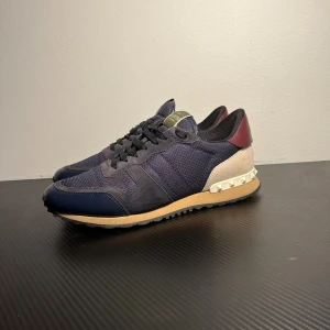 Valentino Rockrunner sneakers blåa  - Snygga Valentino Rockrunner sneakers i mörkblå mesh och mocka med detaljer i beige och vinrött. Sulan är beige med ikoniska pyramidnitar på hälen. Skorna har rund tå, snörning och platt sula. Perfekt för dig som gillar exklusiva och trendiga sneakers.