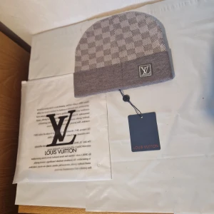 Grå rutig mössa Louis Vuitton - Snygg grå mössa från Louis Vuitton med klassiskt rutigt mönster och LV-logga framtill. Mössan har en ribbad kant och är tillverkad i mjukt material som håller dig varm och stilren. Perfekt accessoar för dig som gillar exklusiva märken.