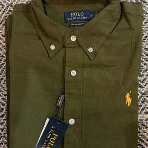 Grön skjorta från Polo Ralph Lauren - Snygg grön skjorta från Polo Ralph Lauren i slim fit med klassisk krage och knappar framtill. Broderad gul logga på bröstet ger en stilren touch. Skjortan är tillverkad i linne, perfekt för dig som gillar en fräsch och modern look.