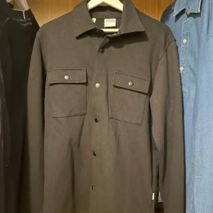 Svart overshirt från Selected Homme - Stilren svart overshirt från Selected Homme med klassisk krage och tryckknappar framtill. Två stora bröstfickor med lock och knappdetaljer ger en clean look. Perfekt lager-på-lager-plagg för dig som gillar minimalistisk stil.