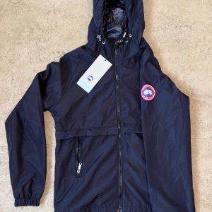Canada Goose Windbreaker  - Snygg svart vindjacka från Canada Goose med huva och dragsko, klassisk logga på ärmen och hel dragkedja framtill. Jackan har en ficka med dragkedja och elastiska muddar vid ärmsluten. Perfekt för blåsiga dagar och enkel att matcha.