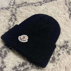 Svart ribbad Moncler mössa - Svart ribbstickad mössa från Moncler med klassisk uppvikt kant och broderad logotyp framtill. Snygg och enkel design som passar perfekt för kalla dagar. Materialet är stickat och mössan har en mörk ton.