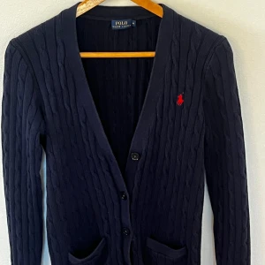 Mörkblå cardigan från Polo Ralph Lauren - Säljer en mörkblå kofta från Polo Ralph Lauren med klassisk kabelstickning, knappar framtill och två fickor. Ikoniska röda loggan broderad på bröstet. Perfekt för lager-på-lager och chill stil. Nypris:2000