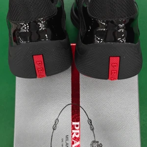 Svarta sneakers från Prada med röd detalj - Säljer ett par svarta Prada sneakers med glansiga paneler och meshdetaljer. Snygg röd logga på hälen och innersulan. Skorna har snörning och en chunky platt sula som ger en modern vibe. Perfekt för dig som gillar exklusiva och stilrena sneakers.