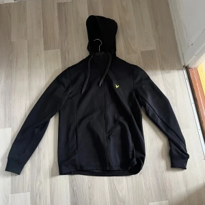 Svart hoodie från Lyle & Scott - Svart hoodie från Lyle & Scott med gul logga på bröstet. Tröjan har huva med snörning, hel dragkedja framtill och långa ärmar. Perfekt för dig som gillar en enkel och stilren look.
