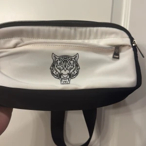 Kenzo svart och vit axelväska med tiger - Snygg axelväska från philipp plein i svart och vitt med ikoniskt tigermotiv framtill. Väskan har justerbart band med philipp plein -logga, dragkedja och mesh-detaljer på baksidan. Perfekt för att bära det viktigaste med stil.