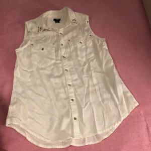 Vit skjorta  - Snygg vit skjorta från H&M i storlek 36, ärmlös modell med krage och guldfärgade nitar på axlarna. Skjortan har två bröstfickor och knappar framtill. Perfekt för dig som gillar detaljer och vill ha en fräsch look.