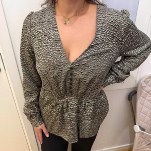 Grå blus med vågigt mönster - Säljer en grå blus med unikt vågigt mönster i svart och vitt. Blusen har v-ringning, långa ärmar och en avslappnad passform. Perfekt för dig som gillar att sticka ut med coola detaljer och mönster.