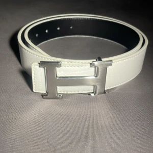 Vitt H-bälte med silver spänne - Säljer ett stilrent vitt H-bälte i skinn med ett ikoniskt H-spänne i silver. Bältet har en klassisk, rak form och är svart på insidan. Perfekt för dig som gillar lyxiga accessoarer med clean look och vill ha något som sticker ut.