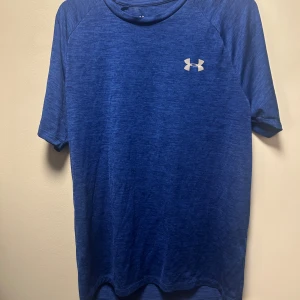 Blå Under Armour t-shirt - Snygg blå t-shirt från Under Armour med diskret logga på bröstet. T-shirten har korta ärmar och är tillverkad i ett lätt och funktionellt material som passar perfekt för träning. Enkel design med rund hals och sportig känsla.