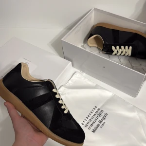 Maison Margiela gats sneakers svart - Maison Margiela gats sneakers i svart skinn och mocka med beige innersula och klassiska vita snören. Snygg kontrast mot den gummifärgade sulan. Ikonisk och stilren modell som passar perfekt till streetwear eller casual outfits. Kom med bud!