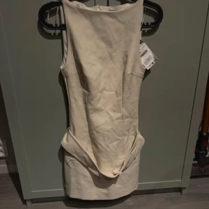 Beige kort klänning med unik detalj - Snygg beige kort klänning från Zara i figurnära modell. Klänningen är ärmlös och har en cool detalj med knappar och en bred panel framtill vid höften. Perfekt för dig som gillar minimalistisk och modern stil.