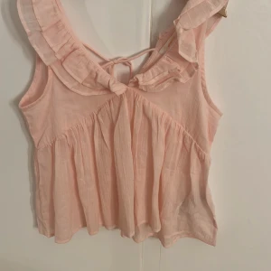 Ljusrosa volangblus med knyt - Supersöt ljusrosa blus med volangdetaljer runt urringningen och knyt i ryggen. Blusen är ärmlös och har en luftig, flowy passform som känns riktigt somrig. Strl 152