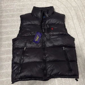 Svart dunväst från Polo Ralph Lauren - Svart dunväst från Polo Ralph Lauren med röd broderad logga på bröstet. Västen har hög krage, tre dragkedjeförsedda fickor och quiltad design. Materialet är glansigt och lätt, perfekt för lager-på-lager. Snygga Polo-detaljer på dragkedjorna.