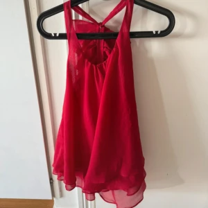 Röd volangig linnetopp med halterneck - Snygg röd linnetopp med halterneck och flera lager volanger framtill. Tunn och luftig modell med lätt transparent tyg och lös passform. Perfekt för dig som vill sticka ut med en färgstark och trendig topp.