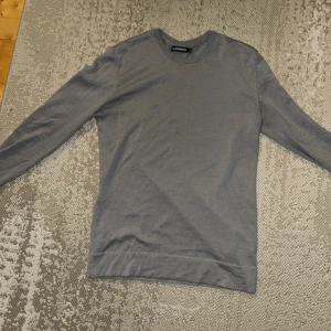 Grå tröja från J.Lindeberg - Snygg grå långärmad tröja från J.Lindeberg. Enkel och stilren design med rund hals och ribbade muddar. Perfekt för dig som gillar minimalistisk stil.