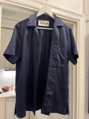 CDLP - Snygg mörkblå skjorta från CDLP med klassisk krage och korta ärmar. Skjortan har knappar framtill och en bröstficka som ger en clean och modern vibe. Perfekt för dig som gillar minimalistisk stil och vill ha något som känns både chill och stilrent.