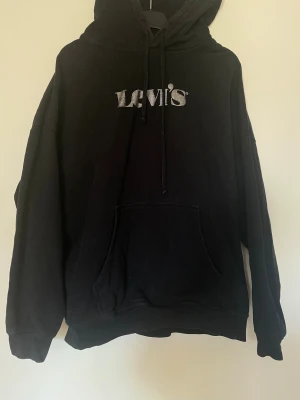 Svart Levi's hoodie med logga - Svart hoodie från Levi's i storlek M med stor vit/skinande logga på bröstet. Tröjan har huva med snörning och en klassisk magficka framtill. Mjuk bomullskänsla och långärmad modell.