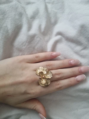 Guldig blomformad statementring - Säljer en stor och unik ring i guldton med en cool blomformad design. Ringen har detaljerade blad och två små kulor i mitten som ger extra karaktär. Perfekt för dig som gillar att sticka ut med dina accessoarer.