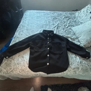Svart overshirt med stora fickor - Svart overshirt med stora bröstfickor och vita knappar framtill. Skjortan har klassisk krage och långärmade ärmar. Materialet ser ut att vara tjockt och mjukt, perfekt för lager-på-lager. Enkel och clean design som funkar till många stilar.