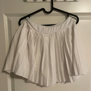 Vit plisserad tennis kjol från SOC - Supersnygg vit plisserad tennis kjol från SOC i storlek S. Kjolen har inbyggda shorts under för extra komfort och rörelsefrihet. Tillverkad i ett tekniskt syntetmaterial som andas och håller dig torr. Perfekt för dig som gillar sportig och fräsch stil.